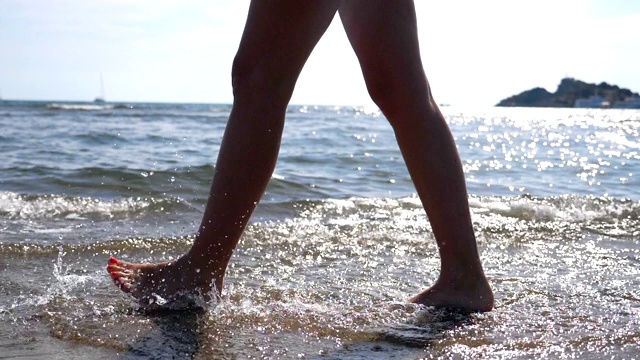 女性的脚走在海水上的海滩与海景和波浪的背景。年轻女子的双腿沿着海岸行走。赤脚女孩在海边漫步。暑假或旅游概念。视频素材