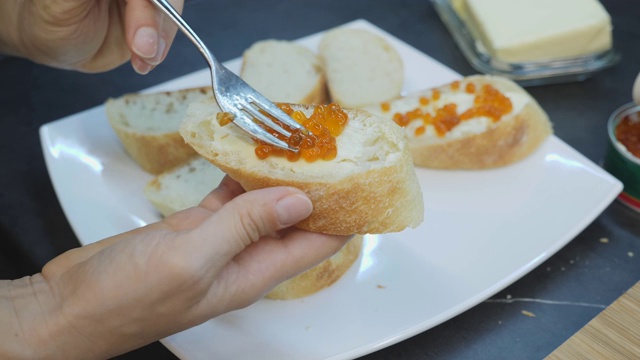 女人把红鱼子酱涂在面包上。用鱼子酱做三明治的特写。家乡菜。将鱼红鱼子酱涂在黄油面包上的工艺视频素材