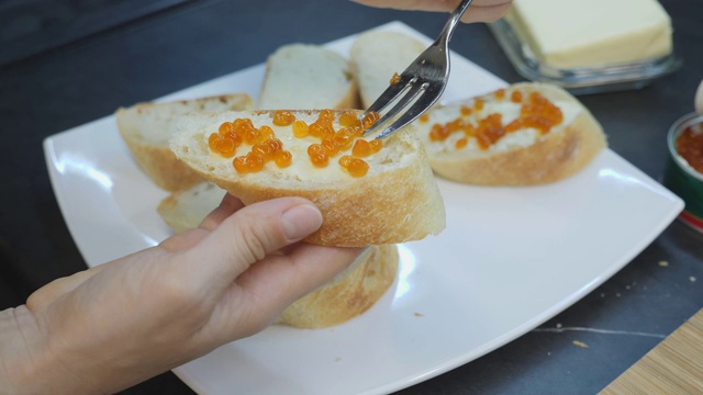 女人用勺子把红鱼子酱涂在面包上。黄油和鱼子酱三明治的特写。家乡菜。女人把红鱼子酱涂在鱼子酱上，放在白色的盘子里视频素材