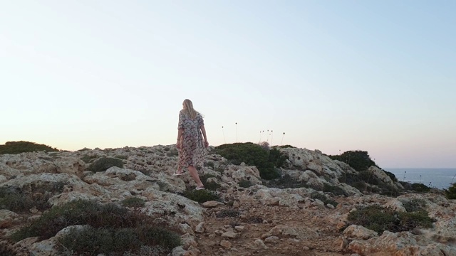 在夕阳西下的格列科角(Cape Greco)，身穿比基尼、身材苗条的金发女子在礁石上漫步，帕雷奥(pareo)在风中飘扬，这是自由和旅行的概念视频素材