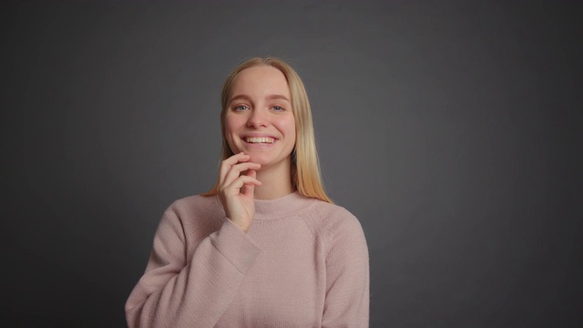 快乐的金发女孩穿着粉红色的毛衣对着灰色的背景微笑视频素材