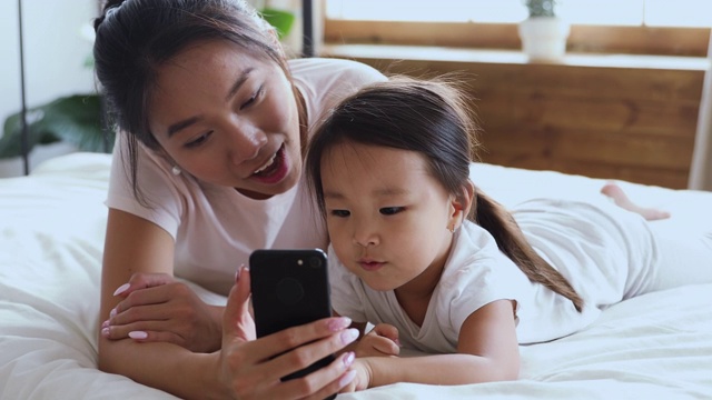 快乐的亚洲母亲使用智能手机和孩子的女儿在床上视频素材