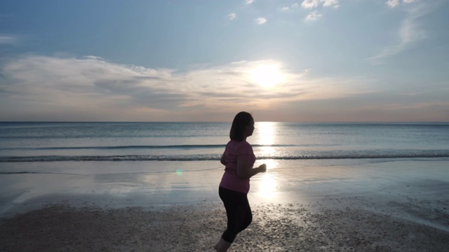 侧视图亚洲身材肥胖的女性正穿着运动服在沙滩上跑步，夕阳西下。健康运动视频素材