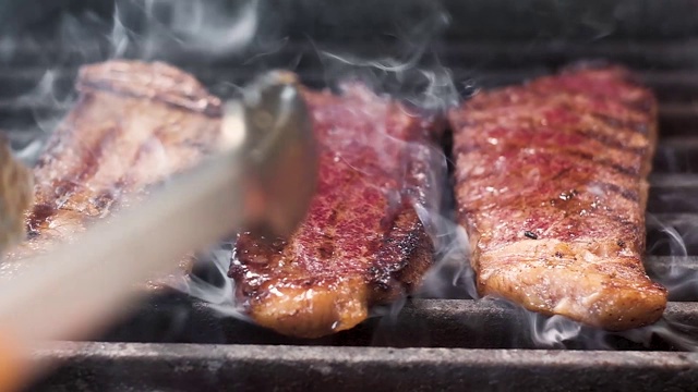 酋长用钳子把三块炸过的肉翻过来。大理石花纹牛肉、牛排。肉上有漂亮的条纹。烤架上美味多汁的食物。慢镜头特写。视频素材