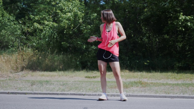 苗条漂亮的女孩戴着耳机，在公园里跳舞，玩得很开心。年轻女子穿着鲜艳的运动服做着奇怪的动作和鬼混。可爱的女孩微笑和大笑。视频素材