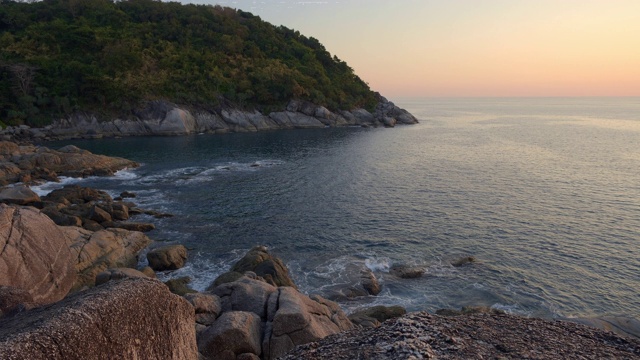 在温暖的夕阳下，岩石海滩上有大而尖锐的石头，山丘和森林视频素材