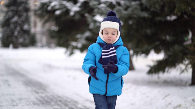 可爱的四岁男孩穿着蓝色的冬装走在下雪的街道上。视频素材