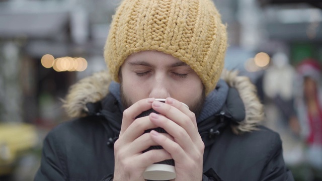 冰冻的年轻白人男子在街上喝热咖啡的特写。一个戴着黄帽子穿着冬大衣的快乐的家伙看着镜头。休闲的生活方式。视频素材