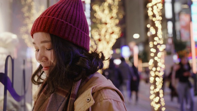 一个亚洲女孩站在商店的橱窗外在一个寒冷的夜晚在城市的圣诞市场，她穿着暖和的衣服。视频素材