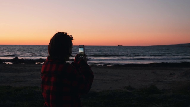 后视图的年轻女性站在海滩上，并使照片和视频的海上日落。女人拍摄美丽的日落。迷人的女人在日落时拍摄大海的照片视频素材