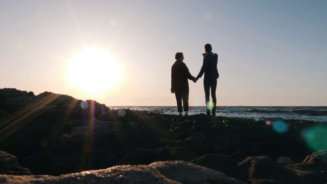 日落时分，一对年轻浪漫的情侣站在海滩上。男人牵着他心爱的女人的手，和她说话，向她展示大海。海滩上一对笑嘻嘻的年轻夫妇的剪影视频素材