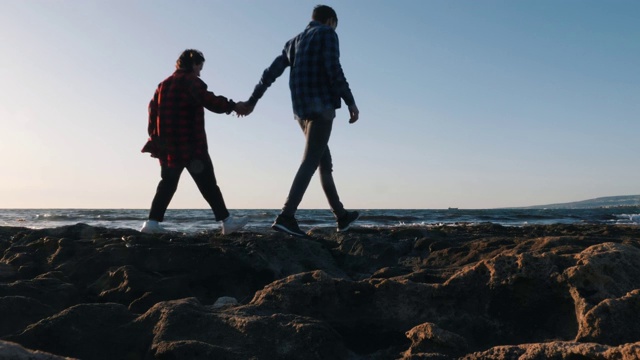 恋爱中的年轻情侣沿着岩石海岸散步。可爱的男孩和他的女孩站在海边拥抱，看着远方。幸福的一对走在一起。男人和女人一起在海上度过时光视频素材