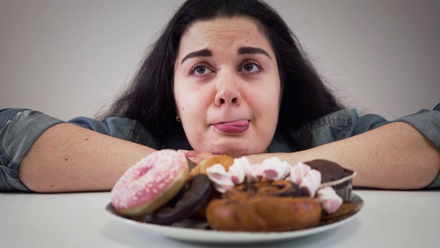 特写脸肥胖的黑发白人女孩坐在盘子前的糖果。饥饿丰满的女人看着美味的甜点和舔嘴唇。超重，健康，不健康的生活方式。视频素材