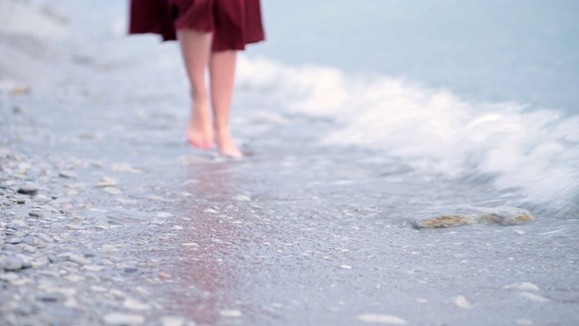 一个穿着红色裙子的年轻女孩的特写腿在潮湿的海岸上翻滚的波浪。从焦点移到焦点。大海的浪花视频素材