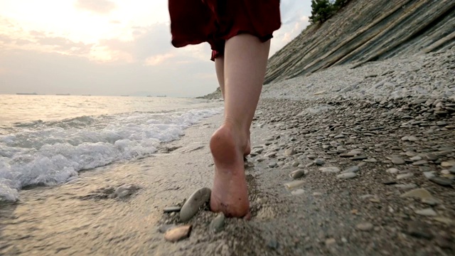 白种女孩沿着海边的石头海岸走向日落与波浪的水视频素材