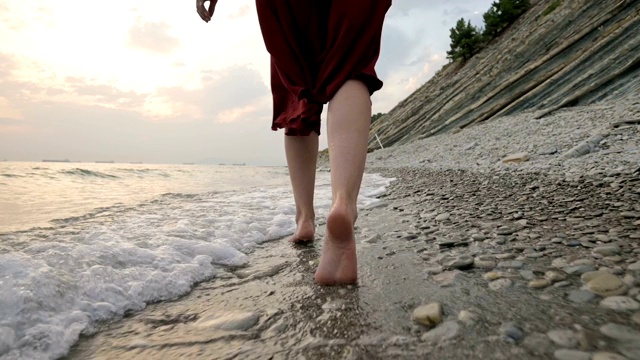 白种女孩沿着海边的石头海岸走向日落与波浪的水视频素材