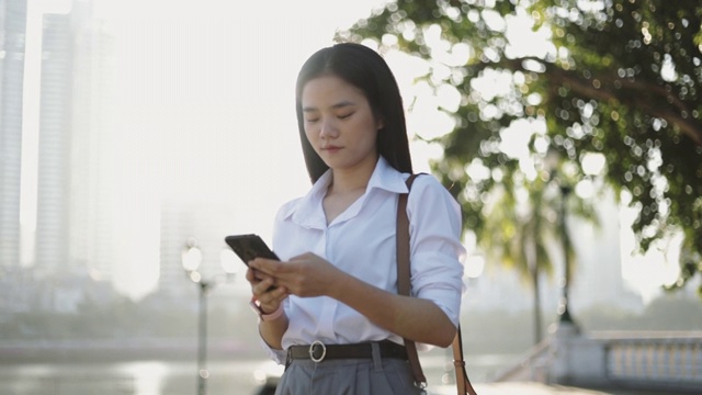 一名身穿白衬衫的亚洲女商人正在公园外用智能手机发送工作信息或电子邮件。视频素材