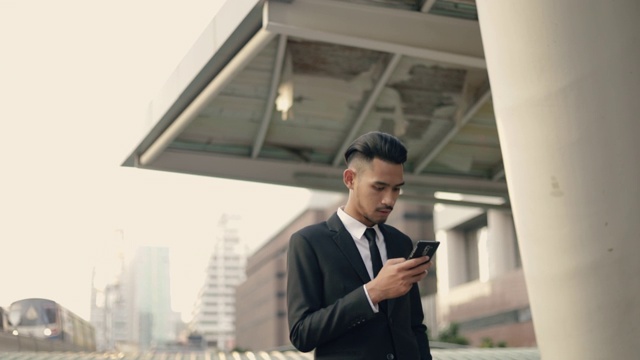 英俊的年轻商人使用智能手机发短信检查电子邮件信息在街道上的城市有空中列车的背景。视频素材