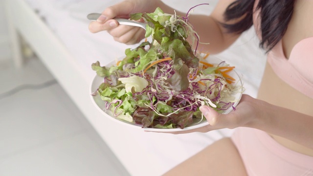 美丽年轻的亚洲女性穿着内衣，身材完美，早餐吃有机蔬菜和沙拉，女孩是减肥与健康的食物，营养和身体护理。视频素材