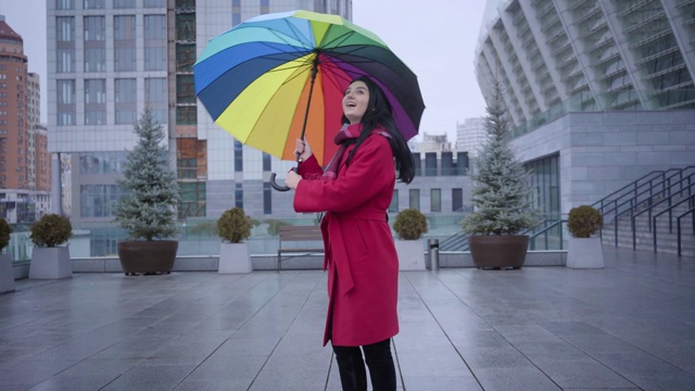 镜头朝积极的年轻女子走来，她拿着五颜六色的雨伞旋转着。可爱开朗的白种女孩与黑色的头发在红色外套享受秋天的雨天在城市。娱乐,生活,快乐。视频素材