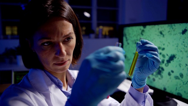 镜头聚焦的女性生物化学家穿着实验服，戴着防护手套，在实验室里仔细检查两个装有液体的试管视频素材