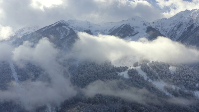 无人机一边空中拍摄冬天山上覆盖的积雪和森林。令人惊叹的冬季景观从无人机雪峰多云的天空背景。白色的薄雾笼罩着山谷和林地视频素材