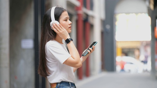 快乐的亚洲女人通过耳机和手持智能手机听音乐时跳舞，亚洲女孩在夏天的户外街道唱歌和跳舞，现代科技和人们的生活理念视频素材