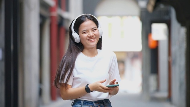 快乐的亚洲女人通过耳机和手持智能手机听音乐时跳舞的慢动作，亚洲女孩在夏天的户外街道上唱歌跳舞，现代科技和人们的生活理念视频素材