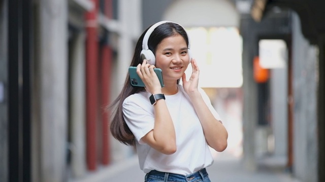 快乐的亚洲女人跳舞时通过耳机，拿着智能手机和看相机，亚洲女孩唱歌和跳舞在户外的街道在夏天视频素材