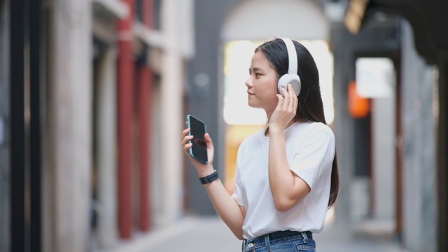 快乐的亚洲女人通过耳机和手持智能手机听音乐时跳舞的慢动作，亚洲女孩在夏天的户外街道上唱歌跳舞，现代科技和人们的生活理念视频素材