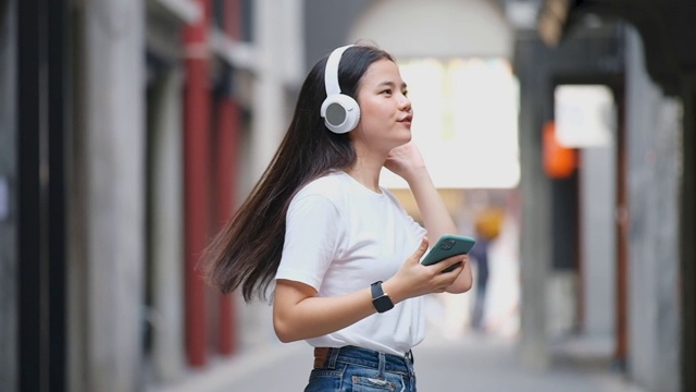 快乐的亚洲女人通过耳机和手持智能手机听音乐时跳舞的慢动作，亚洲女孩在夏天的户外街道上唱歌跳舞，现代科技和人们的生活理念视频素材