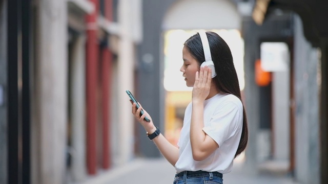 快乐的亚洲女人跳舞时通过耳机和手持智能手机，亚洲女孩唱歌和跳舞在夏天的街头视频素材