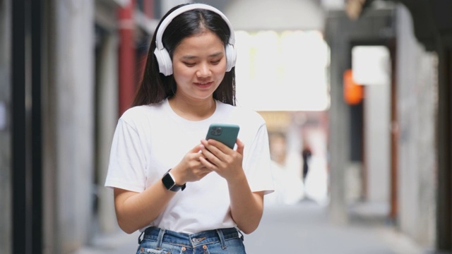 快乐的亚洲女人通过耳机和手持智能手机听音乐时跳舞，亚洲女孩在夏天的户外街道唱歌和跳舞，现代科技和人们的生活理念视频素材