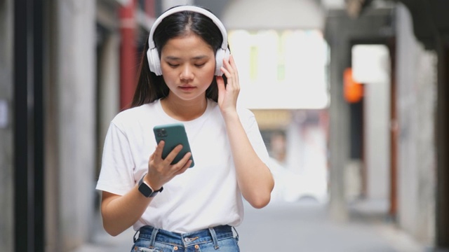 快乐的亚洲女人通过耳机和手持智能手机听音乐时跳舞，亚洲女孩在夏天的户外街道唱歌和跳舞，现代科技和人们的生活理念。视频素材