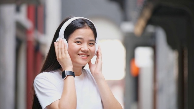 快乐的亚洲女人在耳机里听音乐和看镜头时跳舞的慢动作，亚洲女孩在夏天的户外街道上唱歌跳舞，现代科技和人们的生活理念视频素材