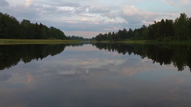 缓慢的运动。在宁静的夏夜，美丽的自然景观。湖岸与绿色的树木和植物反射在水晶干净镜面水面。天空乌云密布。视频素材