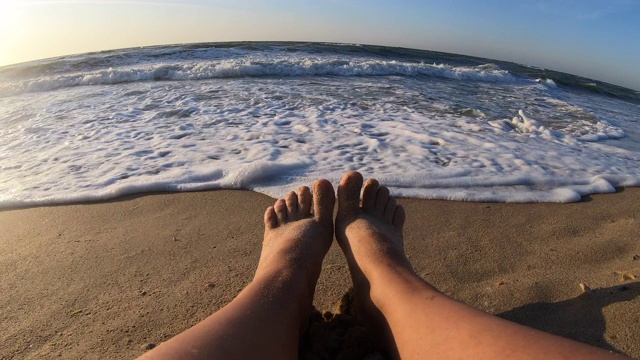 这个女孩坐在海边，沙滩上海浪在她的脚上翻滚视频素材