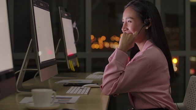 服务电话接线员在无线耳机与麦克风集中在快乐的女人使用电脑在线咨询聊天和微笑在办公室晚上俯瞰博克的灯光交通在城市。视频素材