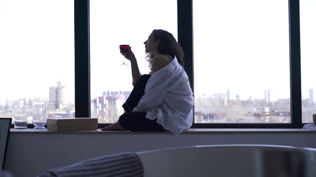 迷人的白人女孩摇晃着红酒杯。年轻的黑发女子坐在窗台上享受着宁静的夜晚。侧视图。视频素材