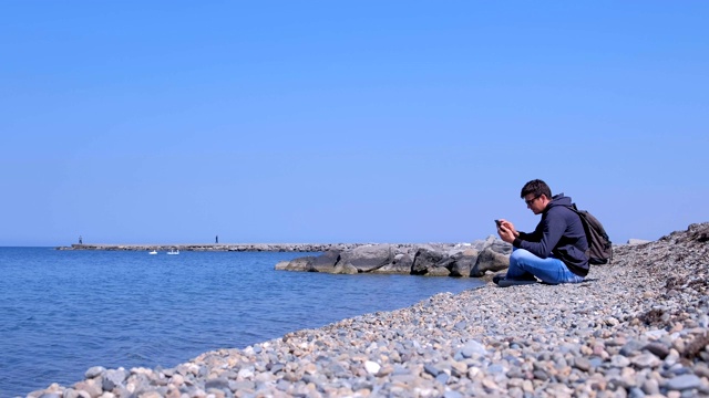 男游客正坐在海石海滩上浏览智能手机，侧视。视频素材