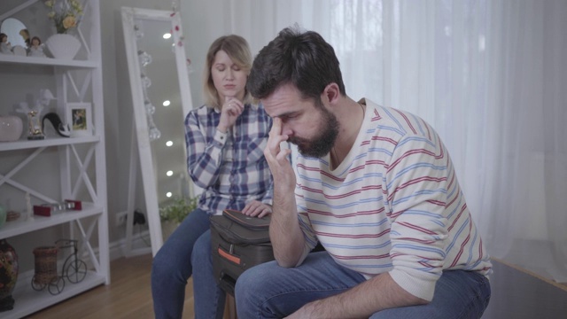 沮丧的白人男子低下头，双手握住它，而女人坐在后面，带着行李箱。恋爱、分手、离婚、分居。视频素材