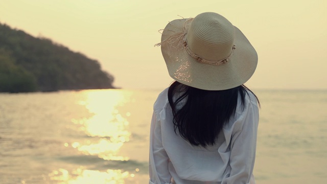 迷人的亚洲女人戴着软呢帽站在海滩上看海景美丽的日落。生活方式旅游度假夏季概念。视频素材