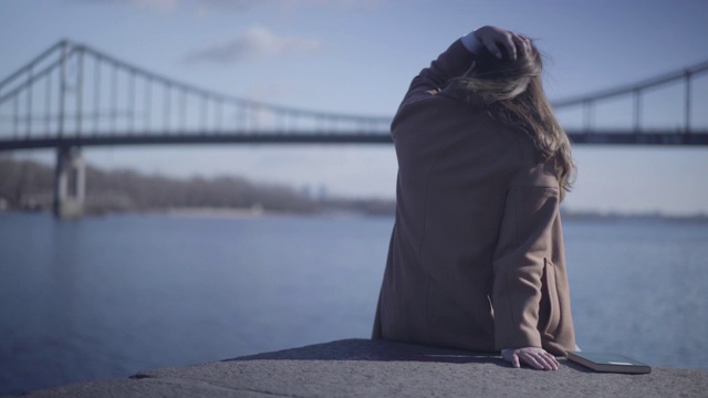 后视图的深色白种女人在秋天的大衣坐在河岸上，看着海浪。平静的小女孩在户外享受阳光。生活方式，休息，休闲，快乐。视频素材