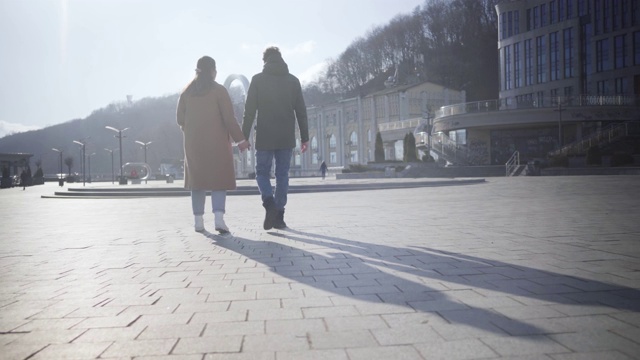 年轻幸福的夫妇漫步在城市广场的宽镜头。白种女人和男人走在阳光明媚的秋日户外的背影。浪漫、约会、爱情、团结。视频素材