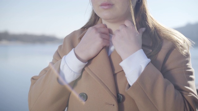 难以辨认的年轻白种女人固定优雅的米色大衣在阳光明媚的秋天一天。画像的胖乎乎的黑发女孩休息在河边的户外。生活方式,休闲。视频素材