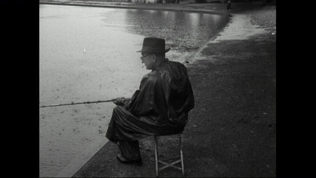 1951年，一名男子在雨中坐在伦敦公园池塘边钓鱼视频素材