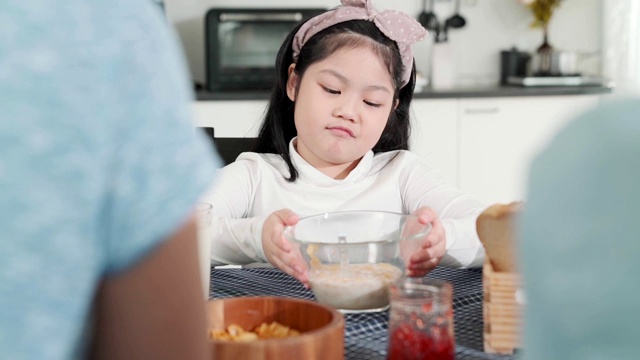 亚洲女性孩子无聊的食物与爸爸妈妈试图说服她吃更多的早餐餐桌上的家庭厨房背景视频素材