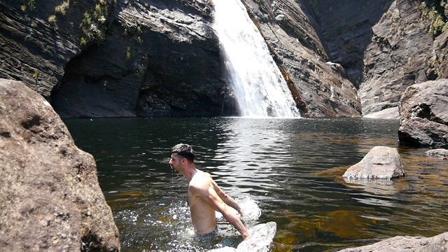 一名男子站在湖中洗脸。男游客在自然的游泳池里休息，享受夏日旅行。奔腾的瀑布倾泻在雨林的垂直岩石悬崖在背景。视频素材