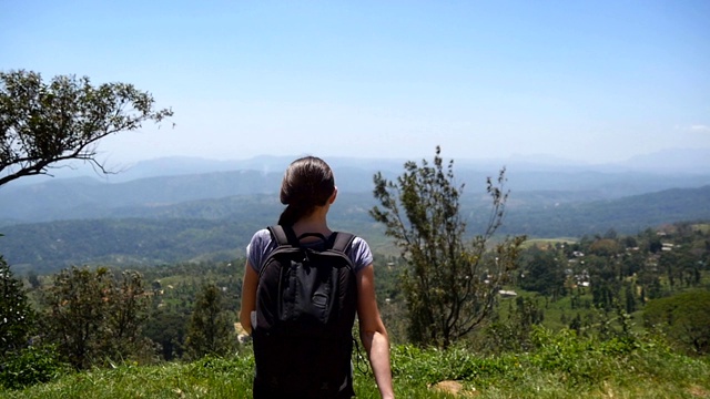 一名背包女游客慢跑到峡谷边缘，举起双手。女性徒步旅行者跑到山顶，伸出手臂。年轻姑娘欣赏美丽如画的风景。后视视频素材