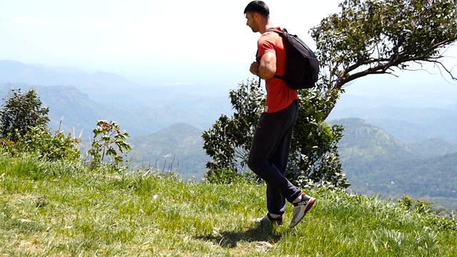 男性疲惫的徒步旅行者与背包走在山间小径和探索地形。年轻的游客走在一条小路上，背景是美丽的山谷。夏季旅游的概念。慢动作视频素材